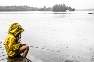 Photo shows a young girl in a yellow rain jacket with the hood up, sitting beside a lake.