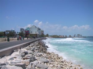 Photo shows a concrete walkway, next to a stoney beach and a blue sea. Buildings in the distance. Sky is blue.