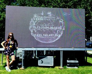 Samantha Mailhot is pictured standing beside a billboard sized projection of the climate clock, showing the time until we reach 1.5 degree of warming, on a sunny summer's day.