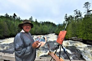 Phil Chadwick, en plein air EcoArtist and Meteorologist, pictured painting outside alongside a river surrounded by trees.