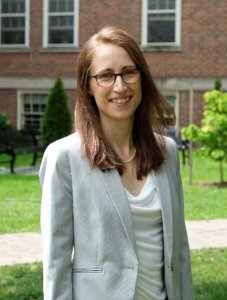 Image shows a photograph of a young woman, straight brown hair and glasses, standing outside a red brick building in the sunshine. Ellen Gute, author of Pollen, Chemistry and Clouds