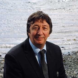 Middle aged Caucasian man with grey-black hair, wearing a black suit and dark grey tie, with an ocean and rocky beach in the background