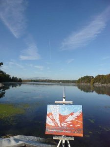 landscape painting in progress on canvas on an easel by the lake. Painting is red, with white marks and blue sky