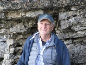 smiling older Caucasian man in blue ball cap and blue outerwear in front of a rockface